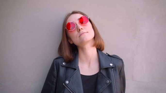 在城市户外，穿着皮夹克和红色太阳镜的年轻酷高加索女孩的特写肖像在镜头前摆姿势