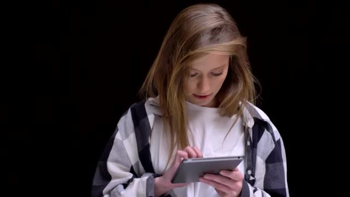 穿着格子衬衫的金发高加索长发女孩的肖像用心地用平板电脑工作，并在黑色背景上对相机微笑。