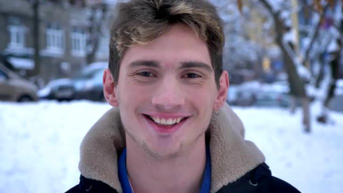 在下雪的冬日里，年轻迷人的白人男性看着相机，微笑着站在外面的特写肖像