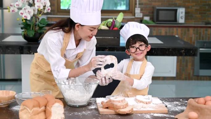 美丽的女人和可爱的亚洲小男孩，戴着眼镜，厨师帽和围裙，在家庭厨房里玩耍和烘焙面包店有趣。自制食物和面