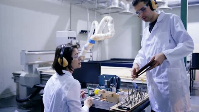 穿着白色外套、防护眼镜和耳罩的工程师向实验室操作激光雕刻机的女同事展示胶合板细节