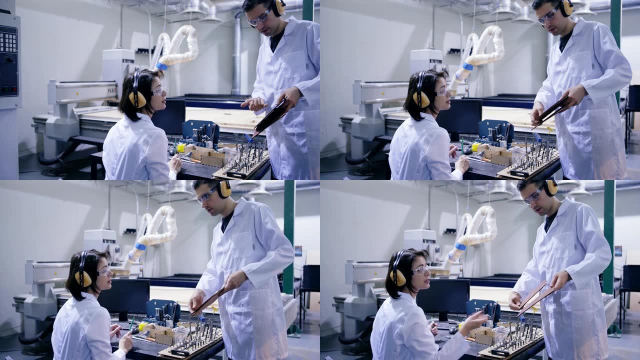穿着白色外套、防护眼镜和耳罩的工程师向实验室操作激光雕刻机的女同事展示胶合板细节