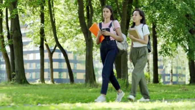 两名亚洲和高加索女大学生的全长跟踪照片，背包，教科书和活页夹在课后穿过绿色公园时交谈