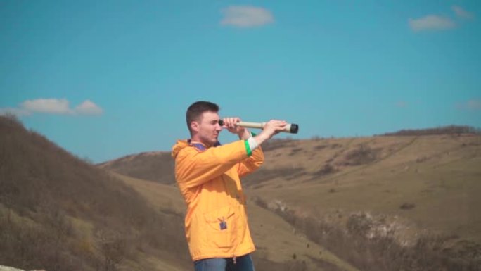 一个穿着黄色夹克和蓝色牛仔裤的年轻人坐在一块岩石上，看着一个望远镜。背景是山脉和天空。