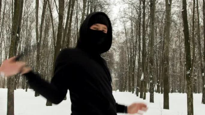 一名武术战士，穿着黑色衣服，脸上戴着面具，在冬天表演拳打双节棍的技巧，白雪覆盖的城市公园在树丛中