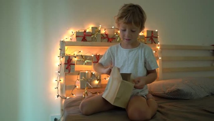 一个小男孩从降临日历上打开礼物的慢动作镜头，该日历挂在用圣诞灯照亮的床上。为圣诞节和新年概念做准备。