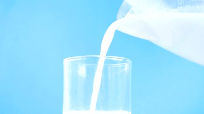 将新鲜牛奶倒入玻璃杯中，慢动作作用于蓝色背景，以促进健康，食物，饮用概念