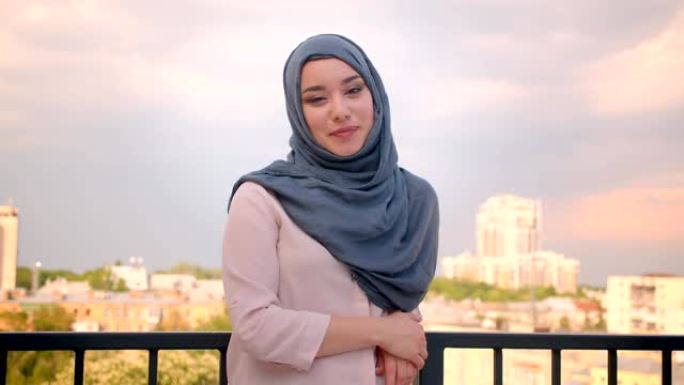 戴着头巾的穆斯林学生的肖像对着镜头微笑，站在阳台上，可以看到城市的美景。