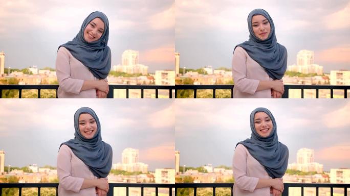 戴着头巾的穆斯林学生的肖像对着镜头微笑，站在阳台上，可以看到城市的美景。