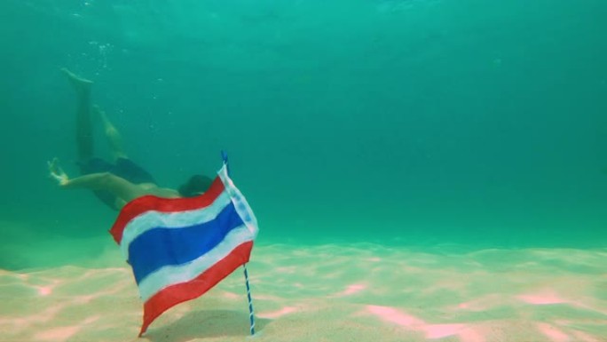 一名男子浮潜潜水并从海底拿走泰国国旗的慢动作镜头。被热带鱼包围的旗帜。泰国旅游概念。泰国的潜水和浮潜