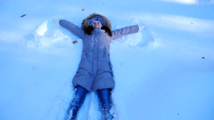 年轻女孩躺在白雪皑皑的草地上，做天使。快乐的女人在雪地里玩耍，享受冬天的时光。开朗的女士在户外玩得开