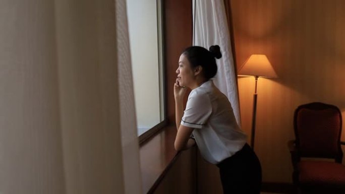 亚洲女性住在酒店房间里。打开窗帘，在房间里用智能手机聊天，看看外面的景色。假期旅行概念。