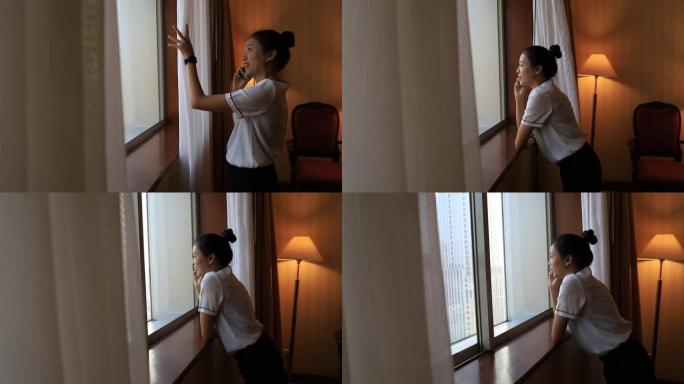 亚洲女性住在酒店房间里。打开窗帘，在房间里用智能手机聊天，看看外面的景色。假期旅行概念。