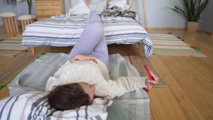 迷人的女孩躺在床边的毯子上，在手机上自拍，鬼混打哈欠。
