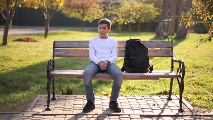 十几岁的男孩穿着白色毛衣坐在长凳上。秋季公园里有背包的帅气男生