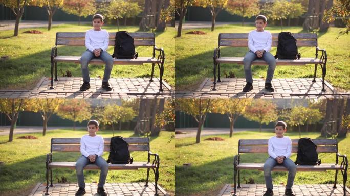 十几岁的男孩穿着白色毛衣坐在长凳上。秋季公园里有背包的帅气男生