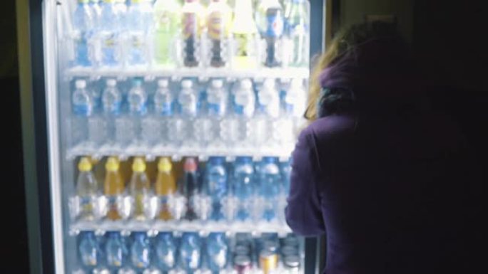 从穿着紫色外套的年轻女子的后面看，从自动售货机上选择饮料。库存镜头。饮料自动售货机出售瓶装水