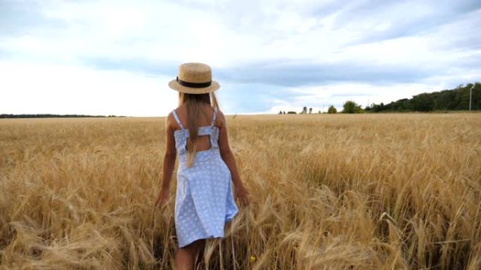 在阴天，跟着戴草帽的小女孩走过麦田。可爱的孩子，长长的金发抚摸着金色的庄稼耳朵。穿着裙子的小孩在大麦