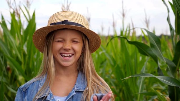 戴着草帽的滑稽小女孩看着镜头，在玉米田上笑着捂着脸。快乐的小孩玩她长长的金发和拉直她的草帽的肖像。