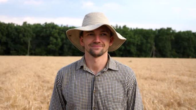 戴着帽子的快乐微笑的农民在麦田模糊的背景下看着相机的特写镜头。英俊的年轻农艺师站在大麦草地上的肖像。
