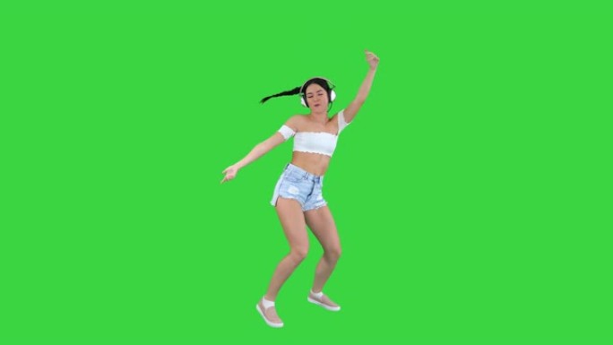 高加索女性模特在耳机中跳跃，表达快乐的情绪听绿色屏幕上的音乐，色键
