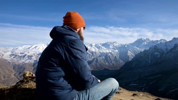 戴着墨镜和帽子的大胡子旅行者的肖像坐在山峦背景下的岩石上