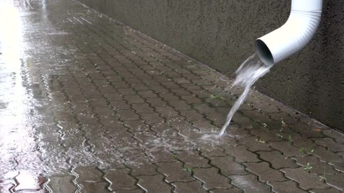 在城市下雨时，水从白色的排水管倾泻到人行道上。特写。地面视图。4K。