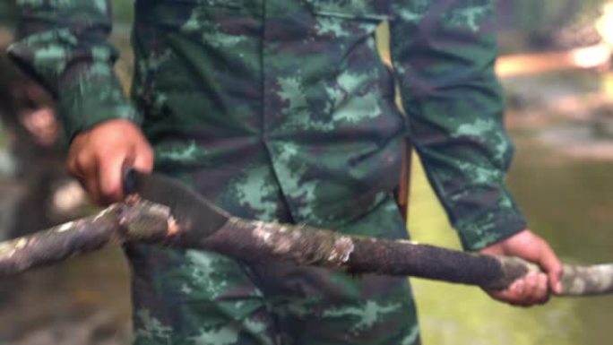 身着迷彩服的士兵用露营刀砍树枝在溪边的森林露营区生火的4K中拍。森林露营用柴火营火。