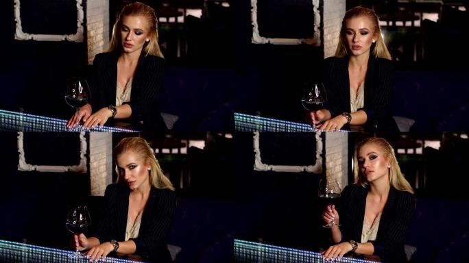 一个年轻性感的金发女郎晚上坐在酒吧，端着一杯红酒。