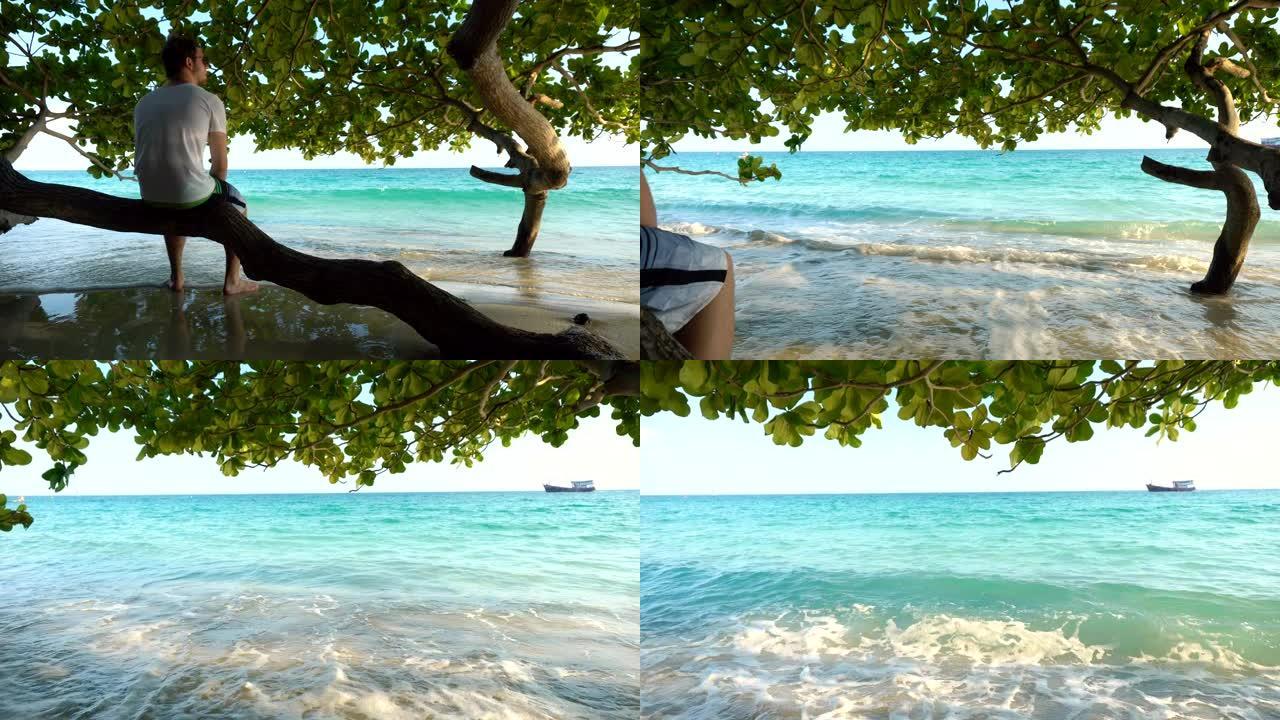 男子坐在热带沙滩上异国情调的树下的阴影树枝上，而海浪抚摸着脚