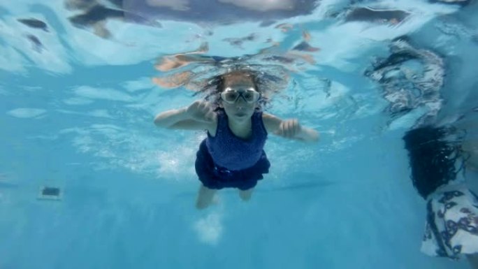 一个小女孩在游泳池里从事水上运动。她戴着泳镜在水下游泳，看着相机。特写。底视图。慢动作。4K。