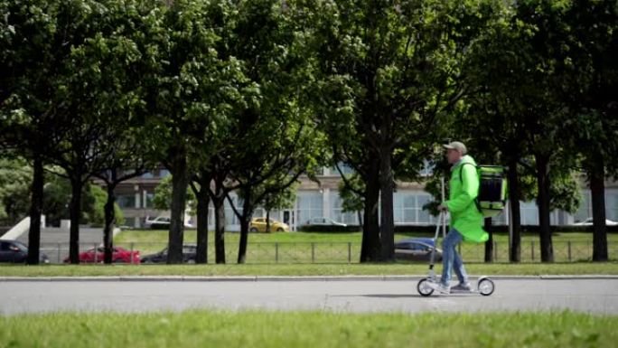 身穿绿色制服的年轻食品快递员在慢动作中骑着踏板车沿着城市街道行驶，背后是隔热背包