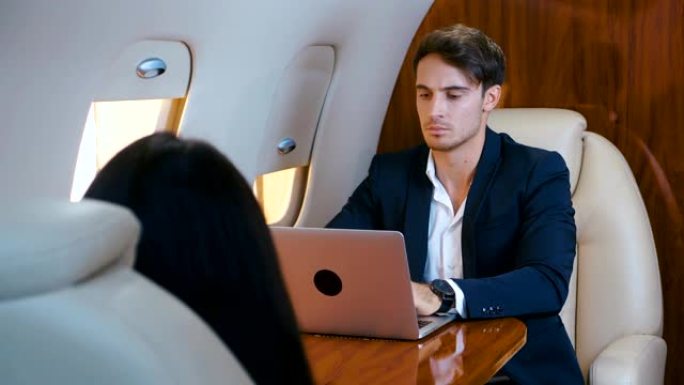 年轻的商人在他的私人飞机上使用笔记本电脑。成功的商业百万富翁乘坐头等舱飞机旅行。穿着古典西装。商人。