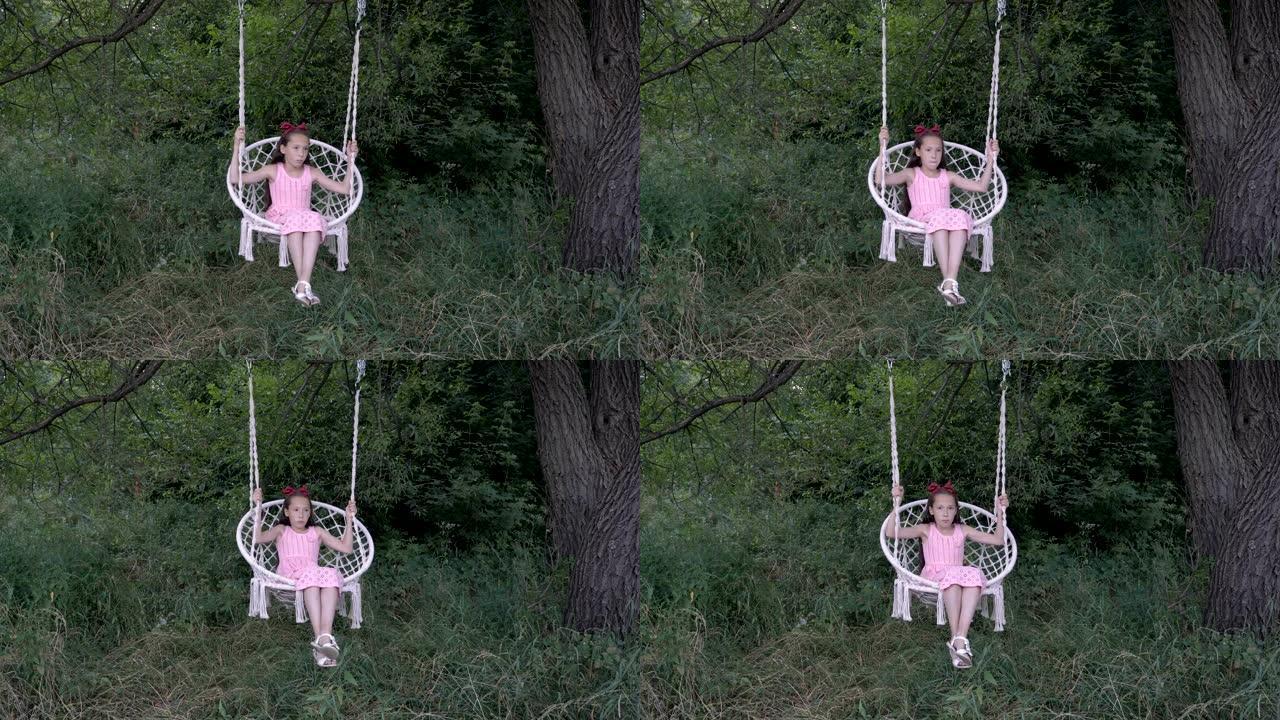 一个沉思的小女孩试图在悬挂在公园树上的悬挂秋千上摆动，背景是绿色的灌木丛。白色秋千上穿着粉色连衣裙的