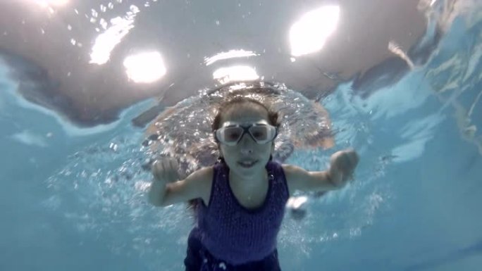 一个孩子，一个小女孩，正在从事水肺潜水。她戴着泳镜在游泳池水下游泳，看着相机微笑。特写。底视图。慢动