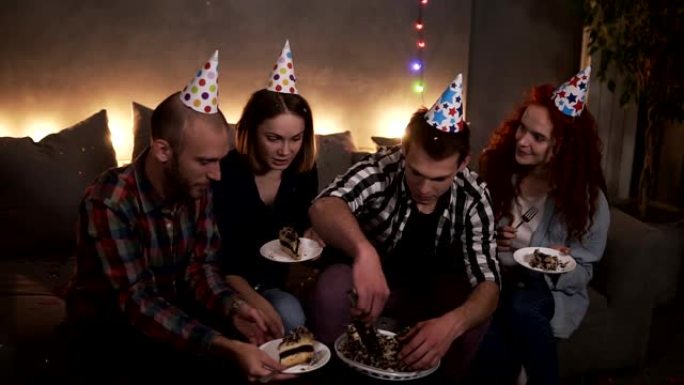 积极的家伙在黑暗中，装饰着花环灯的房间坐在沙发上，吃节日的生日蛋糕。生日男孩把美味的蛋糕放在朋友的盘