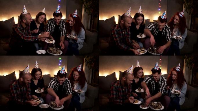 积极的家伙在黑暗中，装饰着花环灯的房间坐在沙发上，吃节日的生日蛋糕。生日男孩把美味的蛋糕放在朋友的盘
