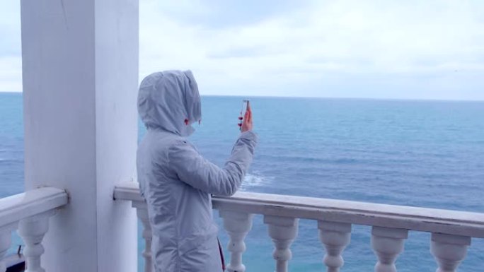 穿着白色夹克的女人在美丽的海景露台上的智能手机上拍摄海浪视频。后视图。