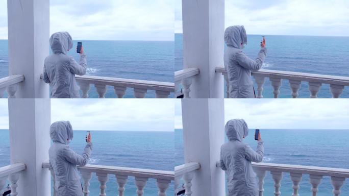 穿着白色夹克的女人在美丽的海景露台上的智能手机上拍摄海浪视频。后视图。