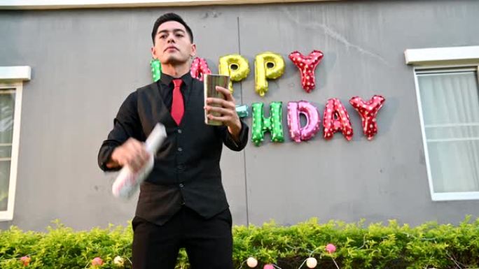 穿着黑色正装和红色领带的迷人英俊的亚洲男性酒保在兼职生日快乐活动中扮演杂耍演员。职业和人们生活方式娱