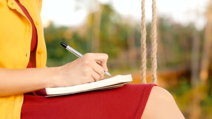 女孩坐在花园里的秋千上，在笔记本上写字，浪漫的年轻女子写日记，写诗自然