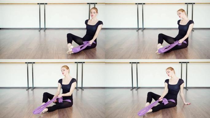 穿着黑色紧身连衣裤和脚鞋的优雅女芭蕾舞演员的跟踪镜头，用弹力带锻炼身体，以改善她坐在芭蕾舞工作室地板
