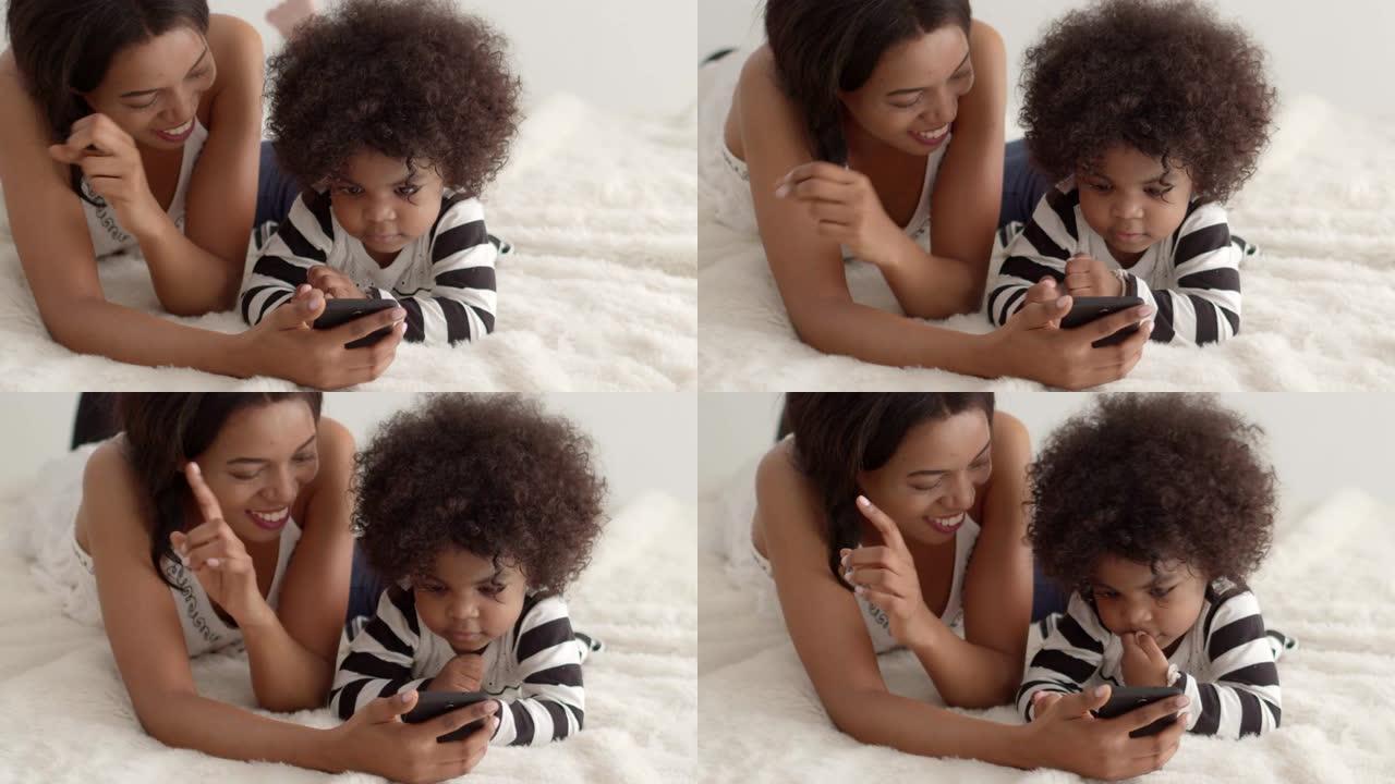 年轻的非洲母亲在手机上看卡通或有趣的歌曲，她的儿子在白色床上。慢动作镜头。