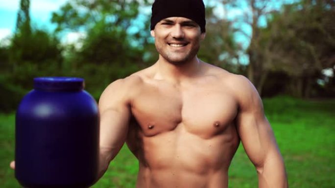 公园里英俊的运动员肖像。肌肉发达的运动员微笑着在镜头前展示一个大蓝色的营养补充品瓶子在健身房和户外艰