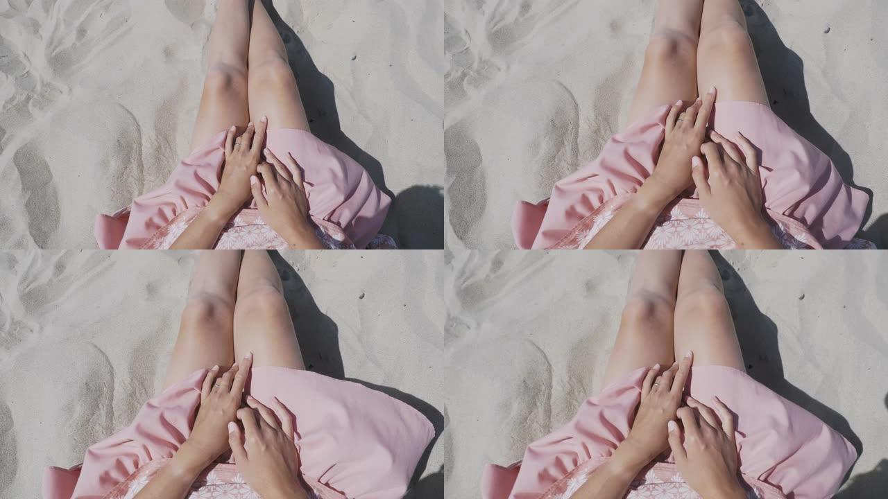 在沙滩上休息的女人。风飘扬的粉色裙子轻盈面料。女人用晒黑的手拿着衣服。从肩膀后面射击。
