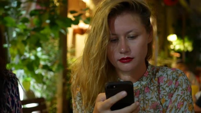 沉思的女孩使用智能手机，写一条消息，聊天。女人在电话里寻找信息而悲伤。独自坐在餐馆里。晚上时间，光线