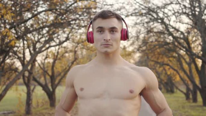 成年白人运动员赤裸上身在秋季公园暖身。强壮的男孩戴着耳机在户外运动前伸展。健康的生活方式的概念。
