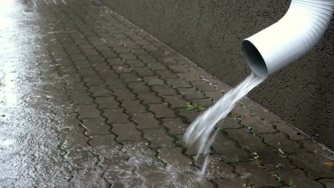 水从建筑物前部的白色排水管倒入铺路板上。阴沉的秋日在城市下雨。特写。侧视图。4K。