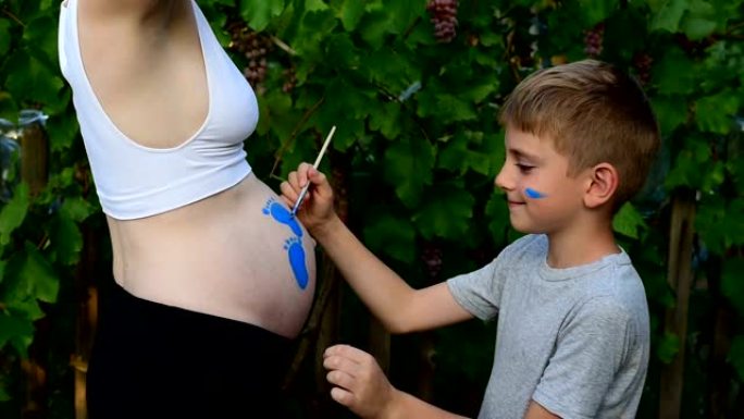 笑男孩用刷子画在他怀孕的妈妈婴儿的脚印的肚子上。怀孕概念。特写