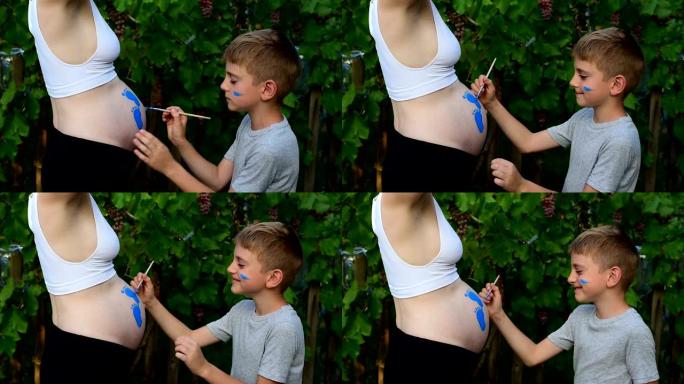 笑男孩用刷子画在他怀孕的妈妈婴儿的脚印的肚子上。怀孕概念。特写